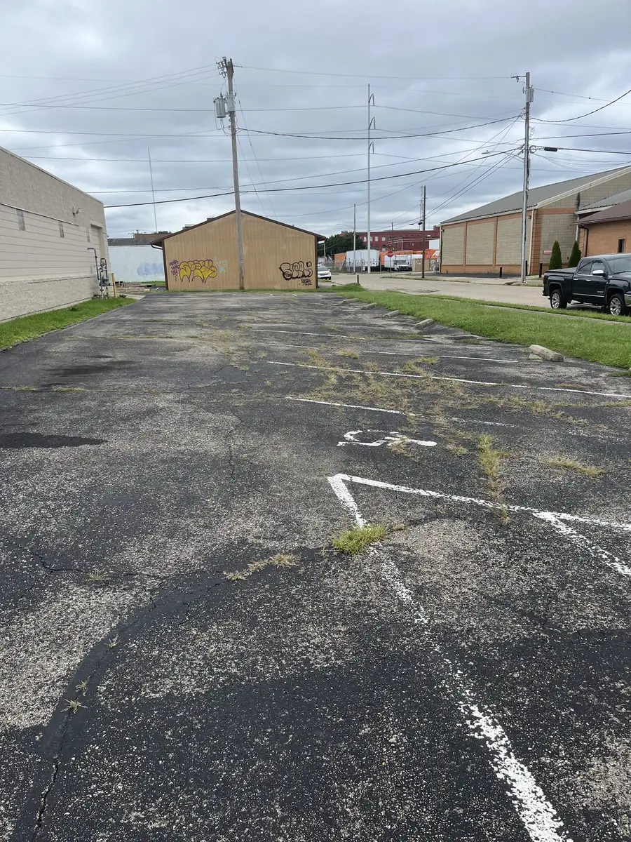 A commercial parking lot before sealcoating, cracked across the surface with weeds growing through the joints and faded white stall markings